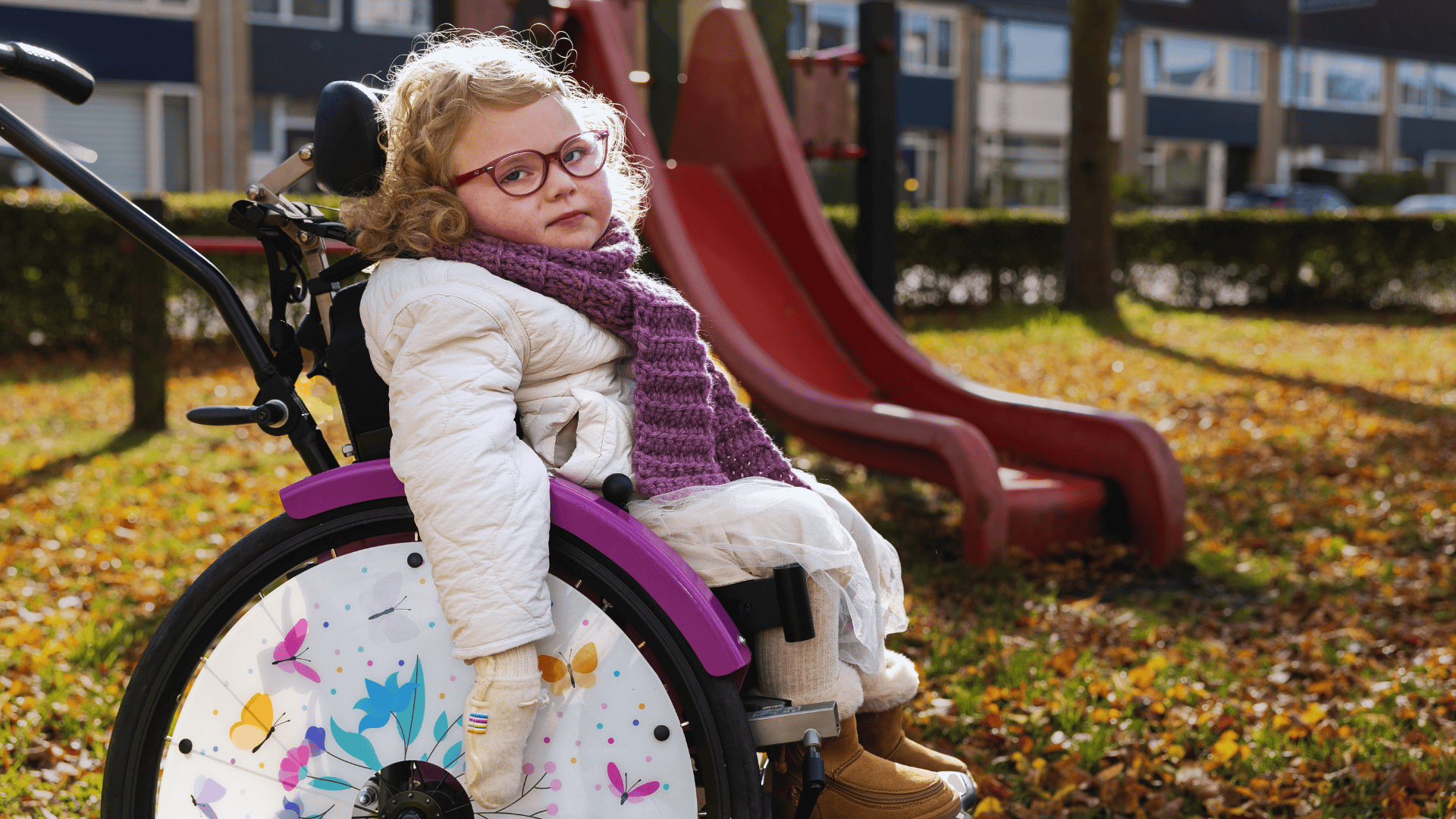 Een jong kind zit in een paarse rolstoel met kleurrijke vlinderdecoratie op het wiel. Het kind draagt een witte jas, een paarse sjaal en bruine laarzen. Op de achtergrond staat een rode glijbaan in een speeltuin, omringd door bomen en herfstbladeren op de grond.
