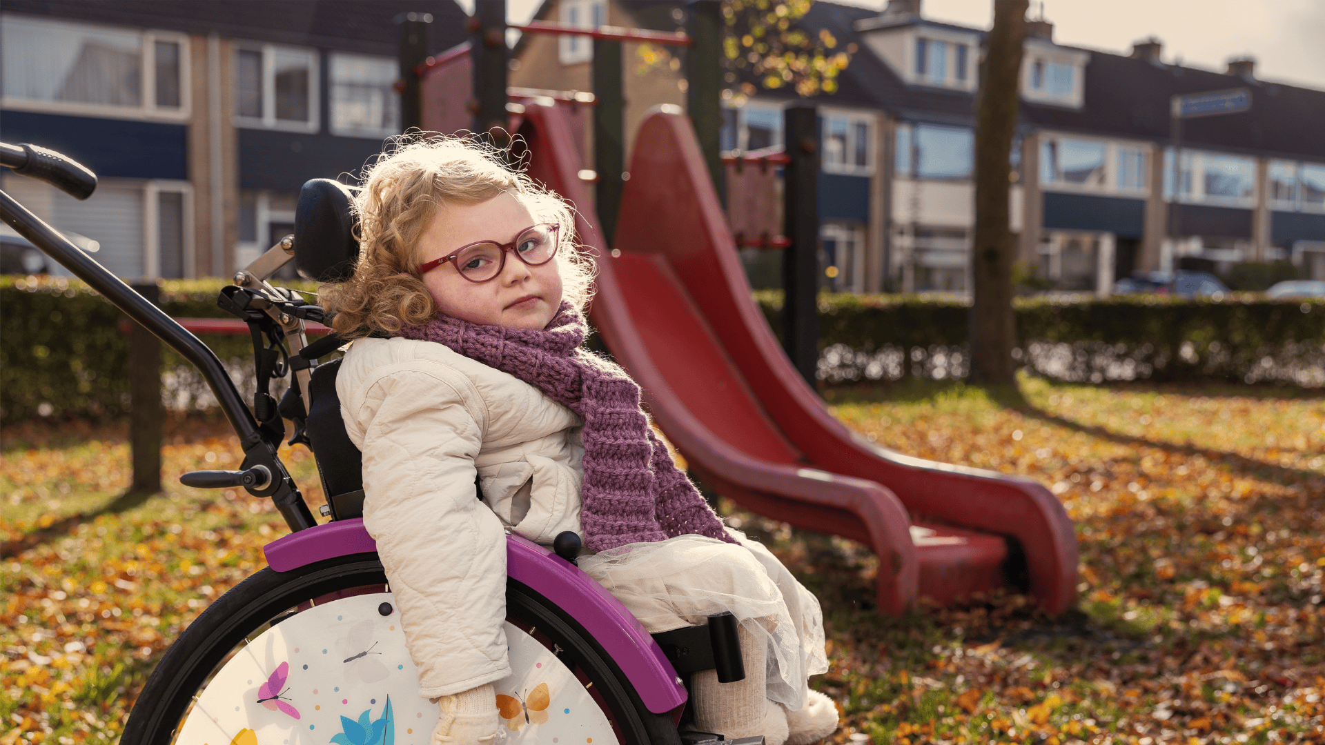 Minthe zit in een paarse rolstoel met kleurrijke vlinderdecoratie op het wiel. Minthe draagt een witte jas, een paarse sjaal en bruine laarzen. Op de achtergrond staat een rode glijbaan in een speeltuin, omringd door bomen en herfstbladeren op de grond.
