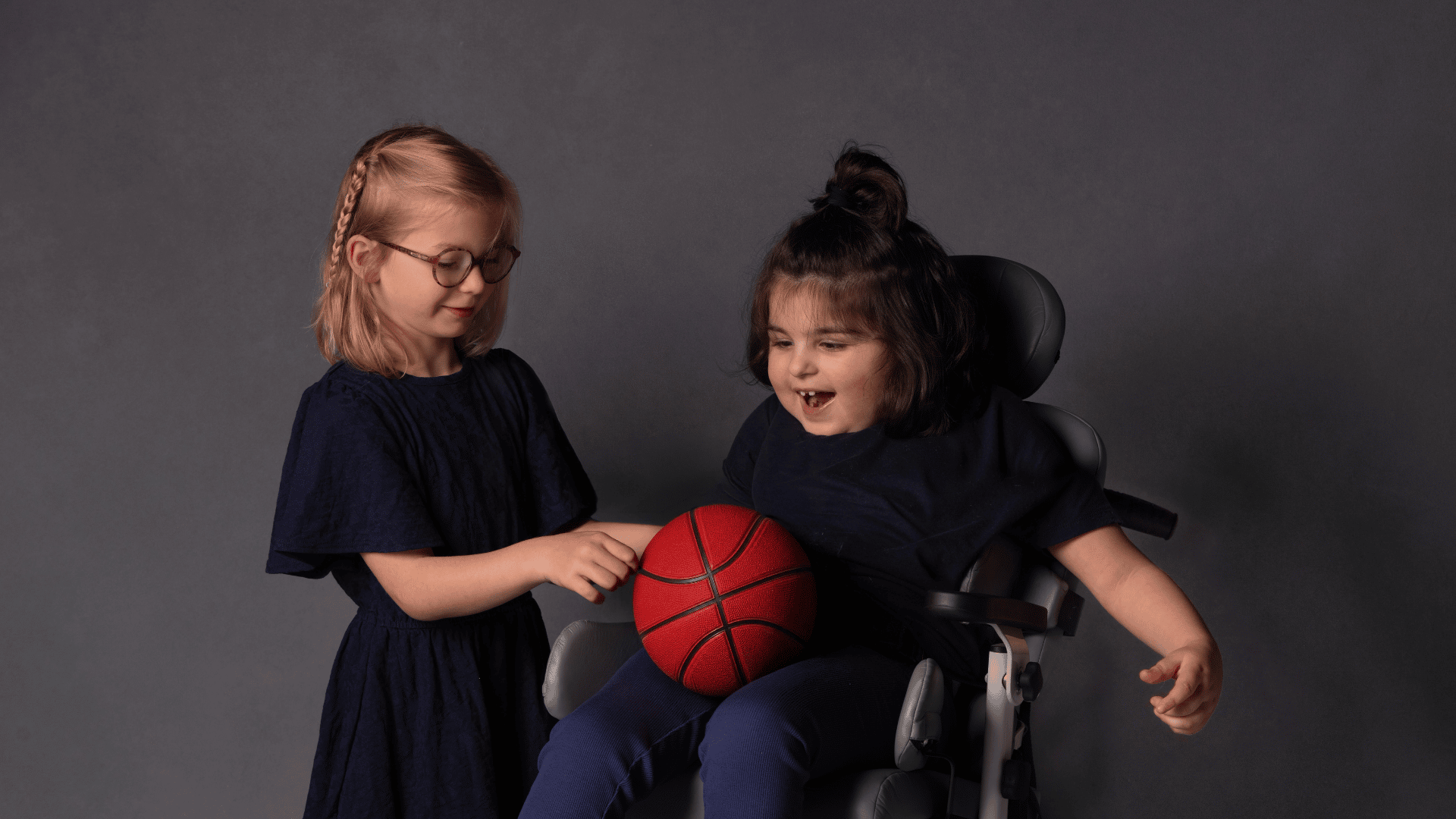 Evi en Sabine staan samen op de foto. Sabine zit in een rolstoel met een bal op haar schoot en Evi kijkt naar de bal