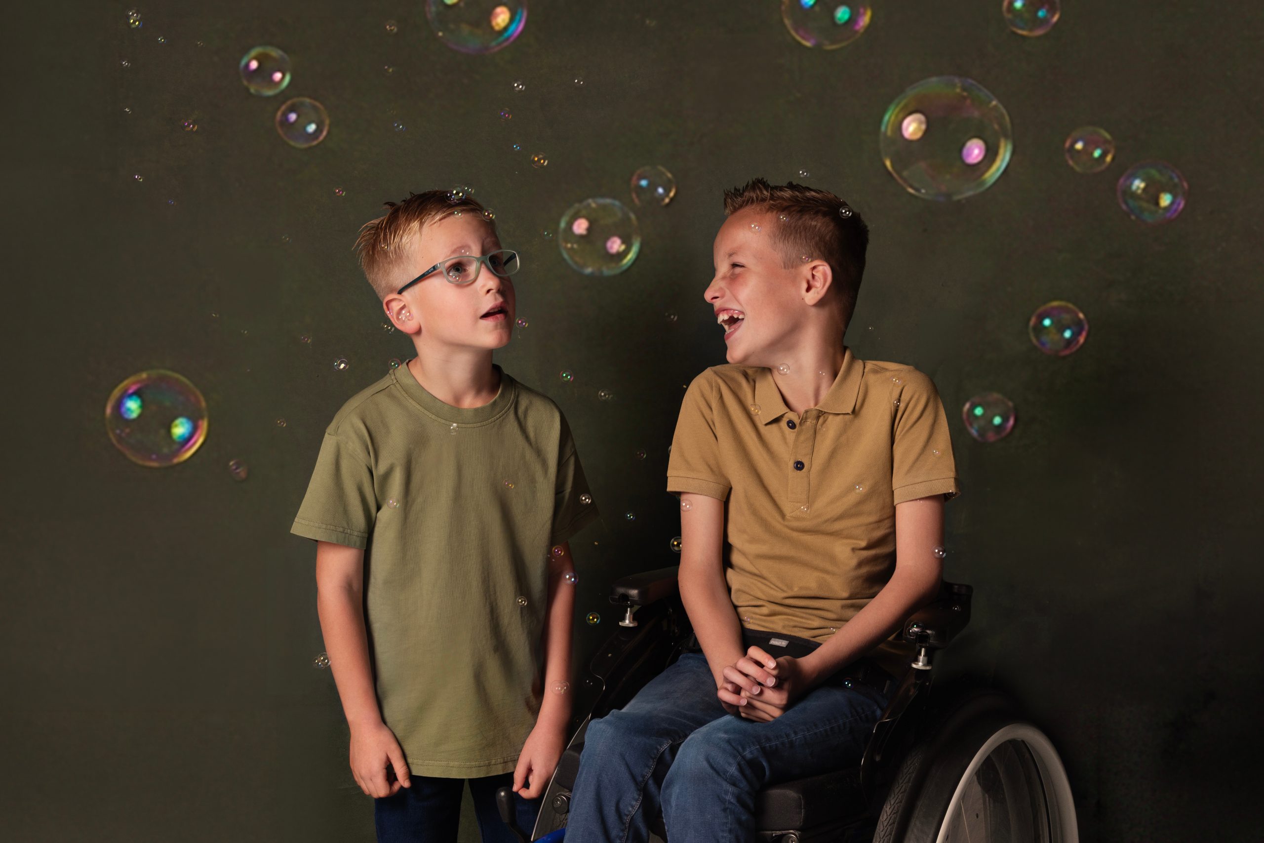 Op de foto zie je Sem en Levi. Sem zit in een rolstoel en lacht. Levi staat links van Sem en kijkt naar de bellen in de lucht.